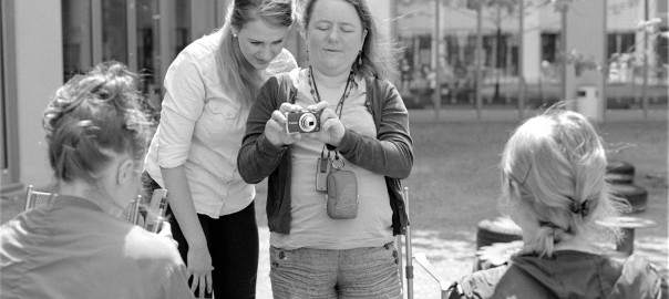 black and white photo of a blind and a sighted woman positioning a camera to photograph another woman in front of them together.